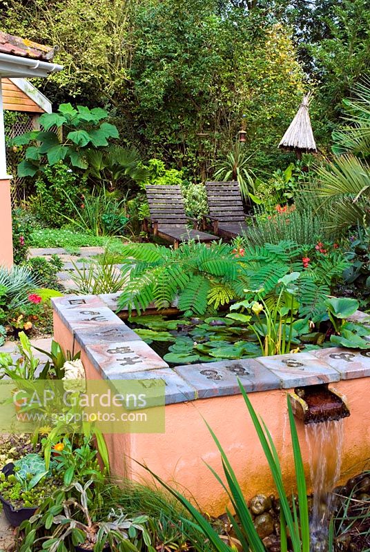 Élément d'eau de piscine peint en terre cuite surélevée avec cascade sur la terrasse d'un jardin de style exotique avec des sièges et des plantes tropicales