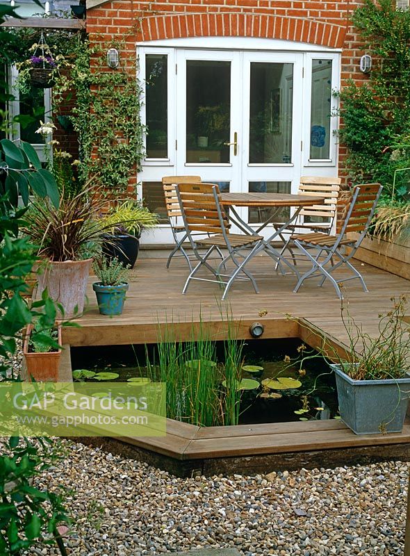 Terrasse en bois surélevé en petite partie gravillonnée, jardin urbain avec étang, table et chaises en bois et pots