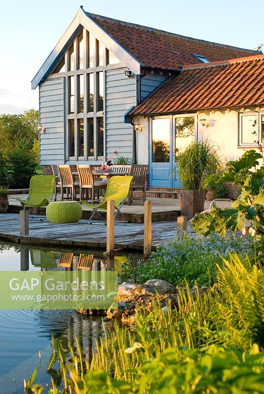 Vue sur l'étang de baignade à la terrasse en bois, à l'extérieur de la salle à manger avec table et chaises et grange convertie avec la chaleur du soleil d'été reflétée sur le bâtiment - Carpe Diem, Bressingham, Norfolk