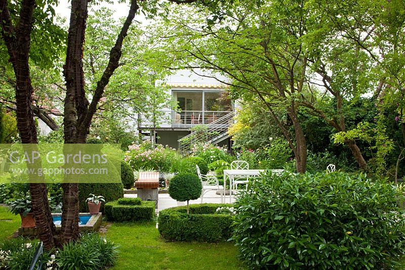 Jardin ombragé avec salon de jardin, topiaire, une haie et un petit bassin d'eau. Les plantes comprennent Buxus, Prunus laurocerasus et Prunus serrula var. tibetica - Allemagne