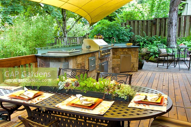 Cuisine extérieure d'été avec grill et évier sur terrasse en bois. Chaises et table en fer forgé avec centre de table en guirlande succulente.
