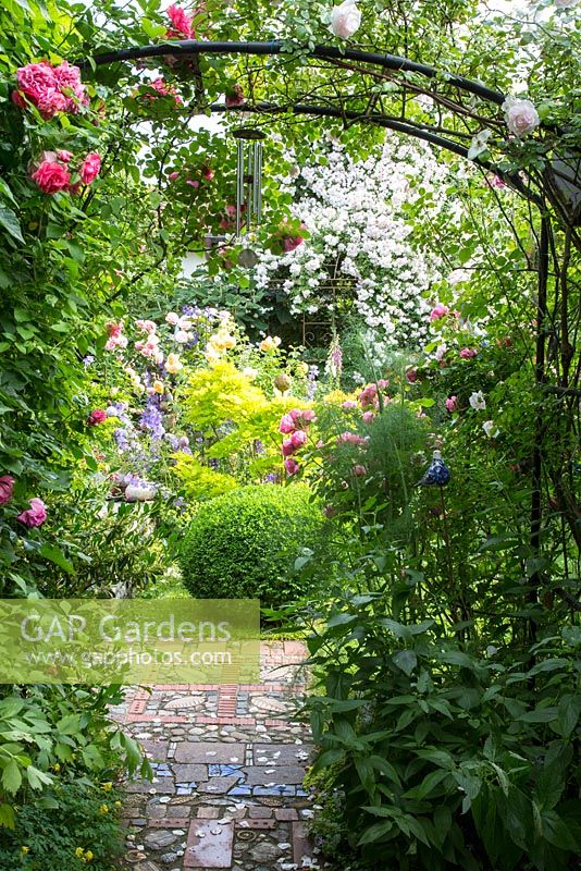 Vu à travers une arche de rose avec des carillons éoliens, une sphère de boîte à côté d'un pavage fou au milieu d'un jardin romantique avec des roses et de l'érable japonais. Les plantes sont Rosa 'Musc himalayen de Paul', 'Rosarium Uetersen' et Buxus