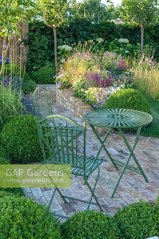Table et chaises en fer forgé sur dallage en briques entourées de balles topiaires - Squire's Garden Centres Urban Oasis - RHS Hampton Court Flower Show 2015