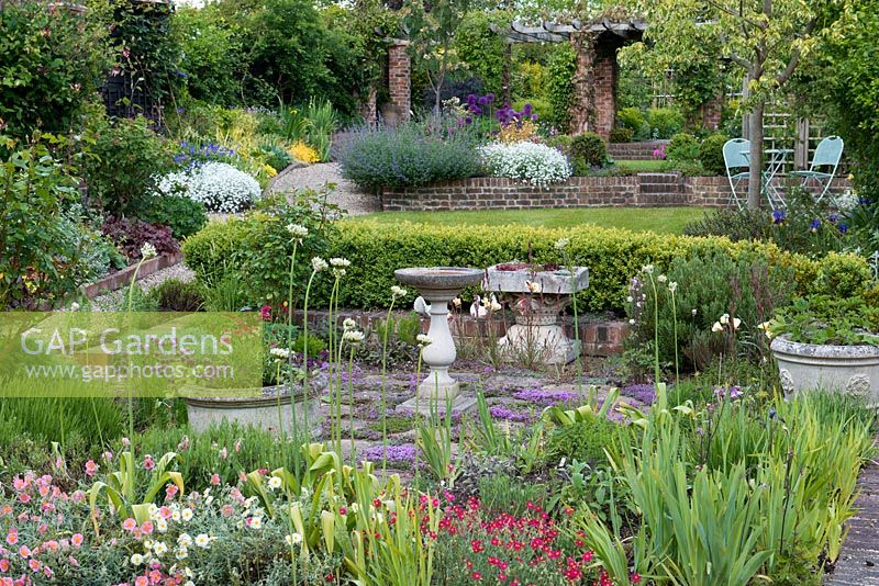 Un jardin à 4 niveaux en terrasses dont un jardin d'herbes aromatiques avec thym intercalé avec dallage, haie de buis, pelouse, coin salon et parterres de fleurs mixtes colorés.