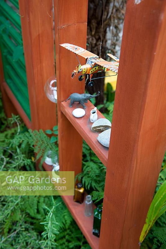 Séparateur de jardin en bois avec niches pour ornements et objets trouvés, y compris un modèle d'avion en étain, des bouteilles et des cailloux. Plantation de feuillage vert.