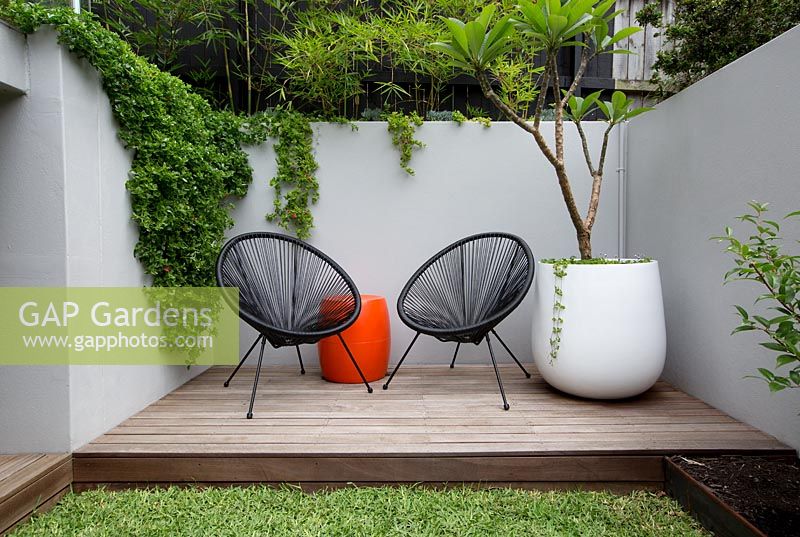 Une terrasse en bois avec deux chaises Acapulco noires, une table à tambour en plastique orange, un pot rond en plastique blanc avec un Plumeria, des frangipaniers devant un mur de soutènement en ciment peint en gris. Avec des plantes succulentes débordant sur le mur et un écran de tisserands élancés en bambou.
