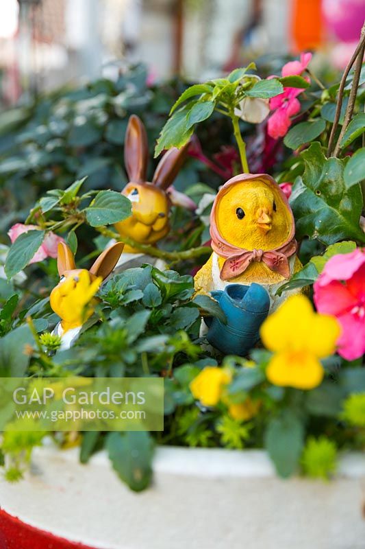 Un pot avec des altos jaunes, des pensées, des Impatiens roses, deux lapins et un poussin avec des visages jaunes et des décorations dans le pot.