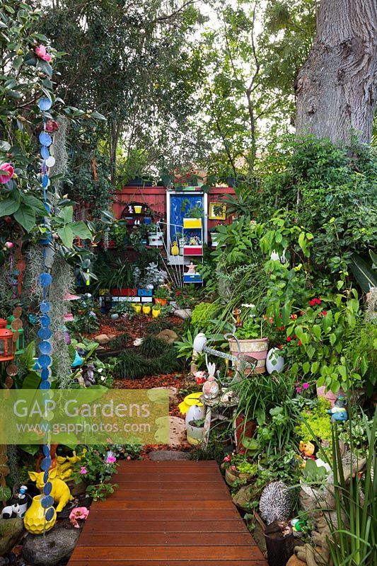 Vue depuis la partie inférieure du jardin jusqu'à la clôture arrière, avec un platelage en bois de forme irrégulière, une collection éclectique de pots, des ornements de jardin, des jouets et des plantes aimant l'ombre.