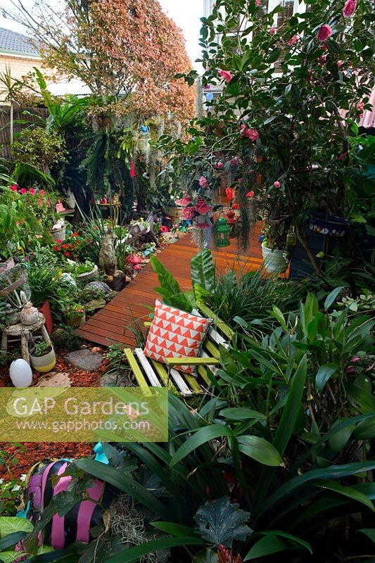 Vue depuis une section surélevée du jardin donnant sur une terrasse en bois de forme irrégulière et un mélange éclectique de pots, d'ornements de jardin, de plantes aimant l'ombre et d'un siège de jardin à lattes avec un coussin à motifs orange.