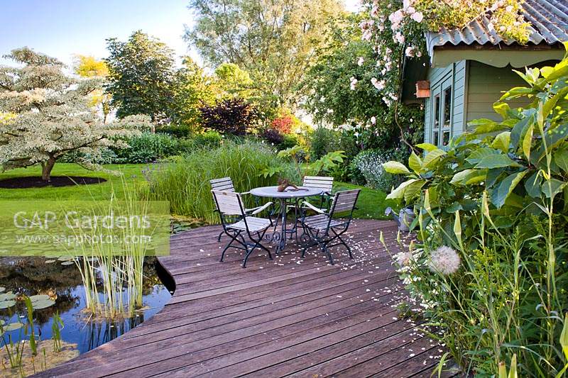 Espace aménagé près d'un étang avec table et chaises, vue sur les parterres de fleurs, la pelouse et les spécimens d'arbres