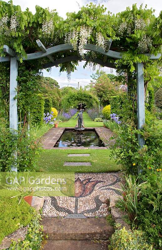 Vue à travers une pergola couverte de glycines jusqu'à un étang rectangulaire avec une sculpture d'une jeune fille se lavant les cheveux par Gerard Laing.