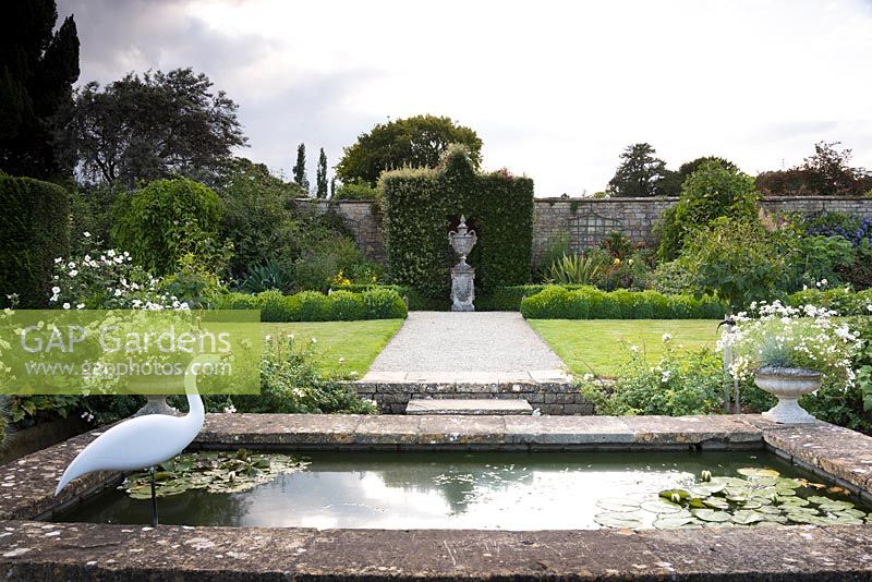 Étang surélevé avec bord pavé avec pots d'Argyranthemum 'Starlight' à chaque coin, vue vers le jardin à la française