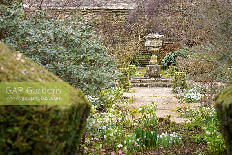 Une urne en pierre au centre du jardin de loisirs, entouré de boîte panachée coupée, perce-neige et hellébores à Rodmarton Manor, Glos, Royaume-Uni.