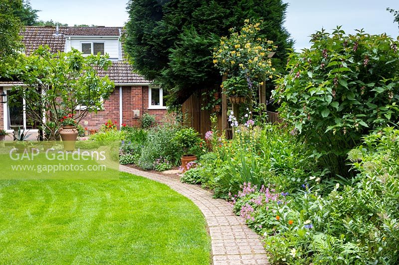 Vue sur jardin avec allée pavée carrée descendant le jardin principal vers la maison, avec parterre ensoleillé à droite.