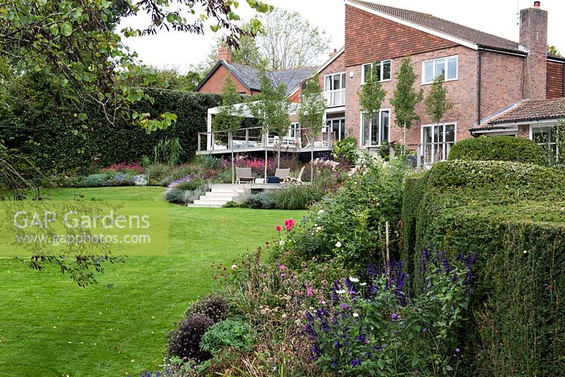 Une conception de maison primée des années 1970, avec des terrasses et des terrasses extérieures contemporaines enveloppées dans des parterres d'automne de plantes à fleurs herbacées et d'herbes ornementales.