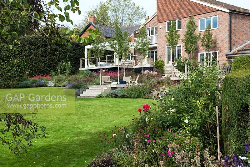 Une conception de maison primée des années 1970, avec des terrasses et des terrasses extérieures contemporaines enveloppées dans des parterres d'automne de plantes à fleurs herbacées et d'herbes ornementales.