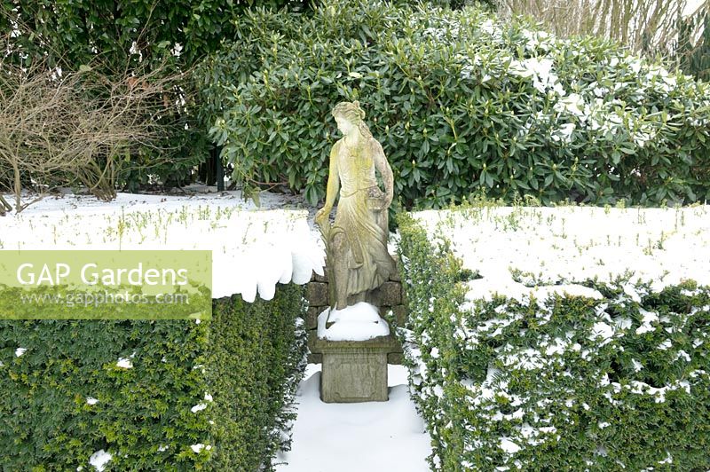 Statue figurative de jardin classique entre les haies couvertes de neige.
