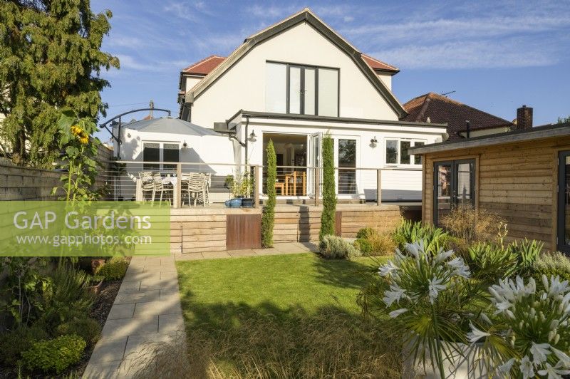 Vue vers maison avec coin salon sur terrasse en bois surélevée et dépendance en bois dans un jardin familial moderne