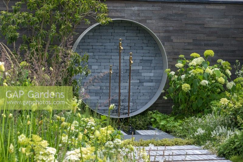 Un endroit pour se retrouver. Hampton Court Flower Festival 2021. Une pièce d'eau circulaire sur le mur recycle les tuyaux et les robinets, flanquée d'Hydrangea arborescens 'Annabelle'.