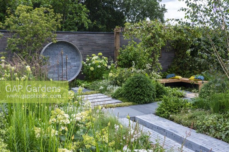 Un endroit pour se retrouver. Hampton Court Flower Festival 2021. Une cour urbaine est plantée dans des tons reposants de gris, vert et blanc, avec une hauteur ajoutée par une pergola drapée de jasmin étoilé. Une fontaine circulaire sur le mur recycle les tuyaux et les robinets.