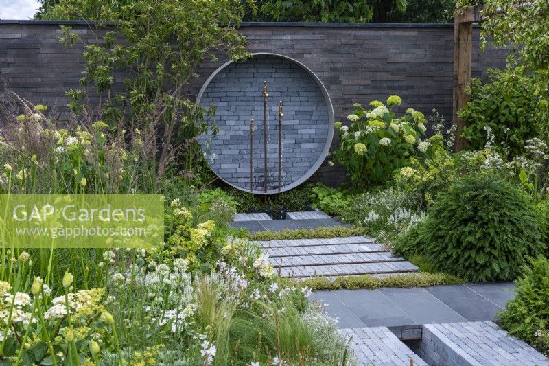 Un endroit pour se retrouver. Hampton Court Flower Festival 2021. Une cour urbaine est plantée dans des tons reposants de gris, vert et blanc. Une fontaine circulaire sur le mur recycle les tuyaux et les robinets.