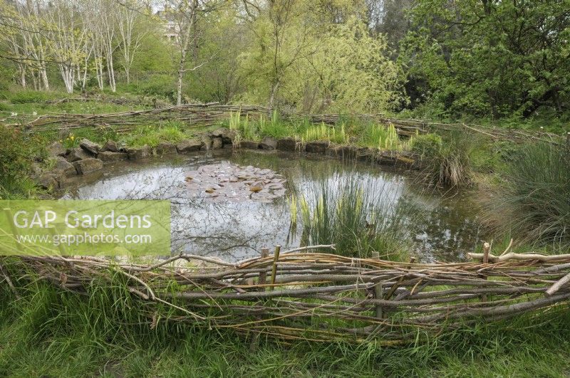 Un étang dans un cadre naturel avec une clôture de platebande extérieure basse faite de branches torsadées et de matériaux en bois.
