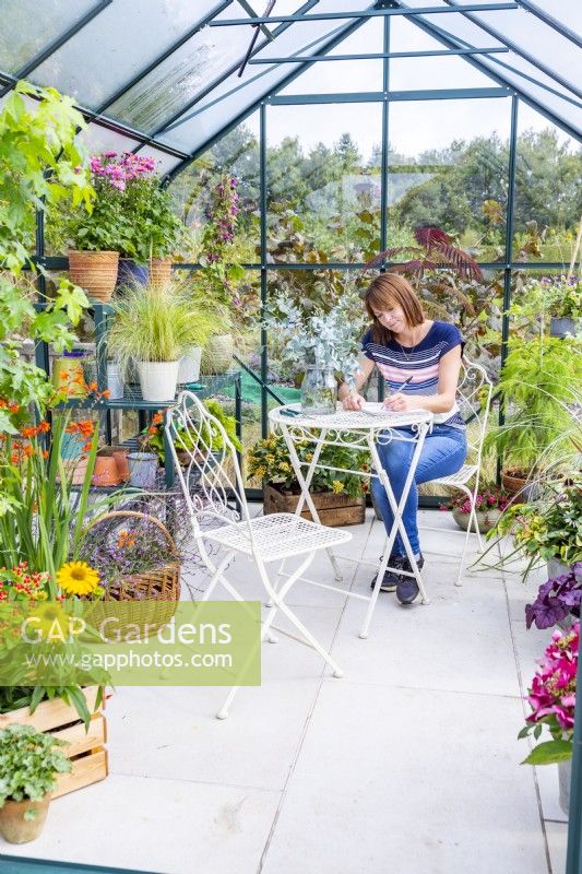 Femme assise à table écrit dans une serre remplie de diverses plantes et pots