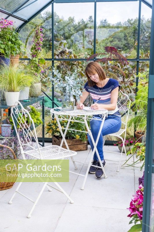 Woman sitting at table writing in a greenhouse filled with various plants and containers 