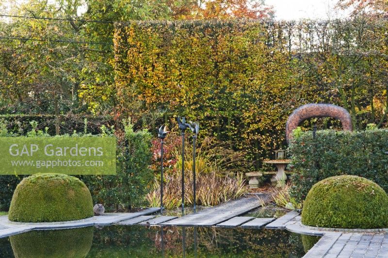 Jardin contemporain avec étang et topiaires de buis, séparé du salon de jardin voisin par des haies de houx, des sculptures ornementales en métal et un accès en pierre à l'espace de détente, adossé à une haute haie de charmes.