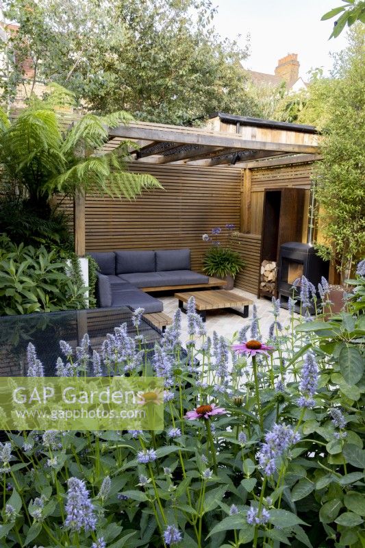 Vue sur un parterre de fleurs herbacées avec Agastache 'Blue Fortune' vers le patio et la pergola en bois contemporaine