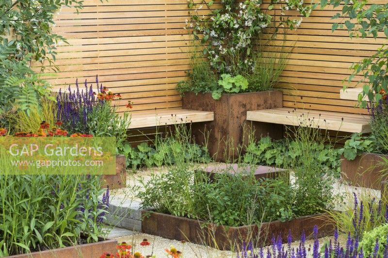 Jardinières en acier rouillé avec plantes vivaces avec Helenium 'Moerheim Beauty' et bancs en bois simples, clôture et pavé en argile - Lunch Break Garden au RHS Hampton Court Palace Garden Festival 2022