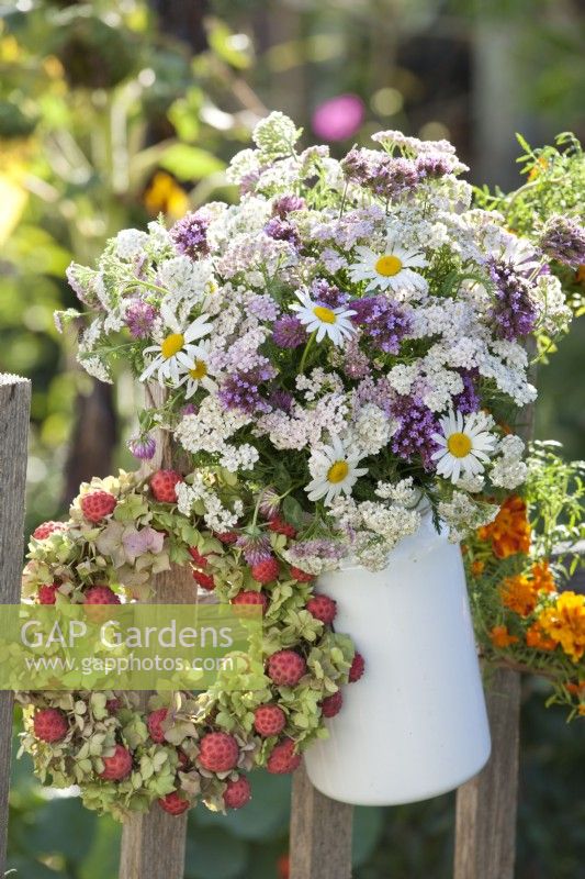 Bouquet of verbena, achillea and daisies in enamel jug and wreath made of hydrangea and Cornus kousa fruits hanging on fence.