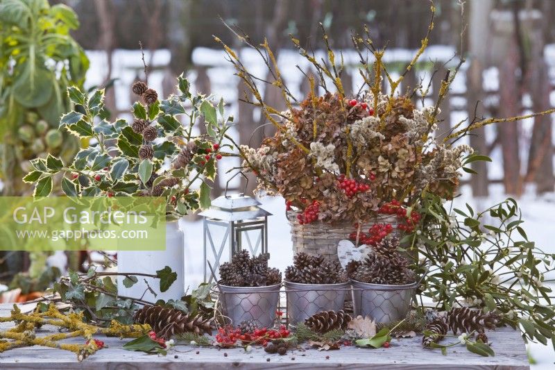 Arrangement hivernal comprenant un bouquet d'Ilex dans un pot de lait, des fleurs d'hortensia séchées et des brindilles de rose de Gueldre avec des baies dans un panier tressé et des cônes.