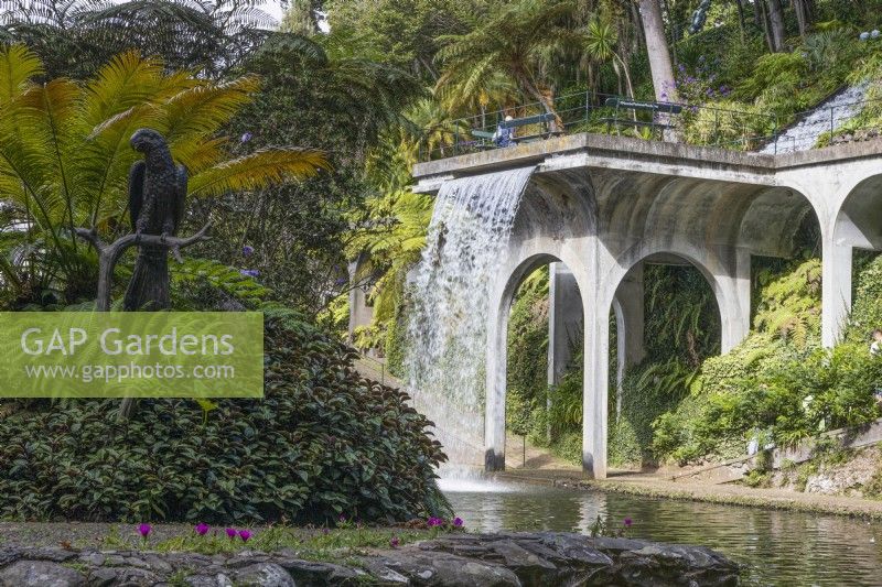 La cascade, conçue par José Berardo, s'écoule depuis un balcon de style viaduc jusqu'au lac Central. Une statue d’oiseau se trouve sur une île avec des plantations tropicales au premier plan. Jardins du Monte Palace, Madère. Août. Été 