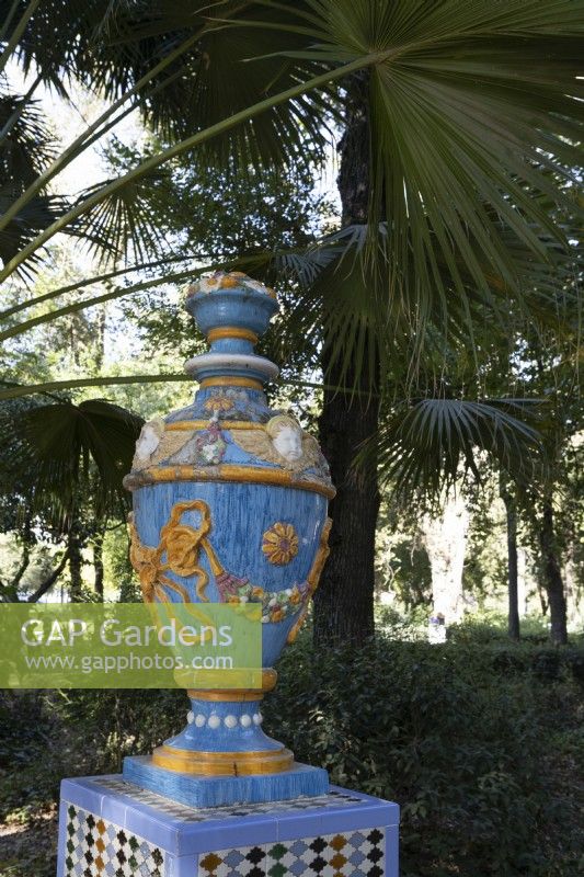 Une urne colorée et vitrée, posée sur un socle. Divers arbustes et arbres sont en arrière-plan. Parque de Maria Luisa, Séville, Espagne. Septembre 