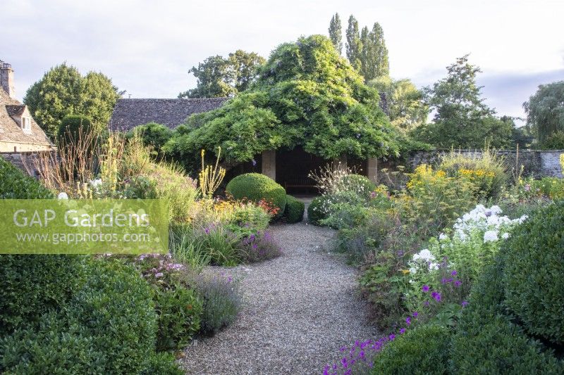 Un chemin de gravier mène au-delà des topiaires et des parterres de fleurs jusqu'à une maison d'été couverte de glycines dans le jardin fleuri du Manoir, Little Compton. 