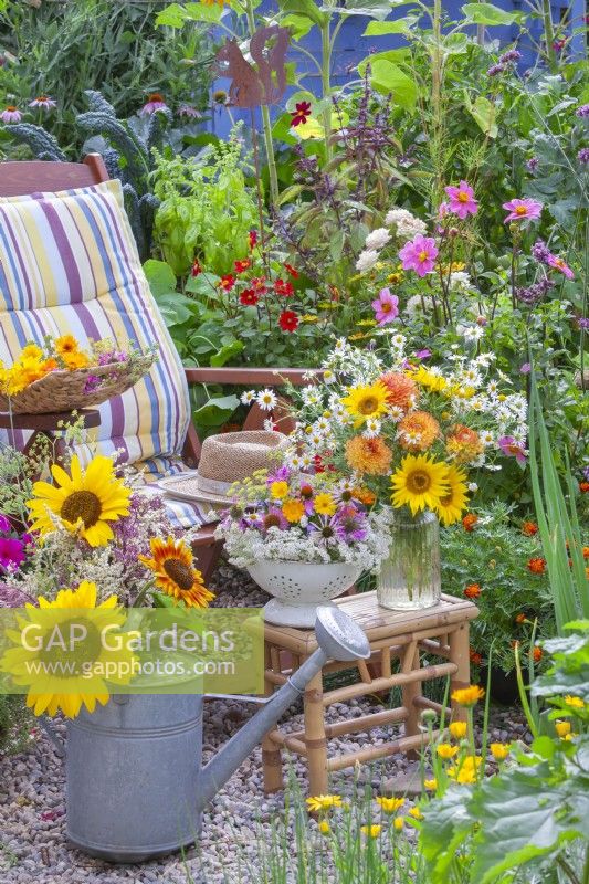Une passoire remplie d'herbes et de plantes médicinales, tandis qu'un arrosoir et un vase en verre contiennent des fleurs d'été, notamment des tournesols, des dahlias et de la camomille. 