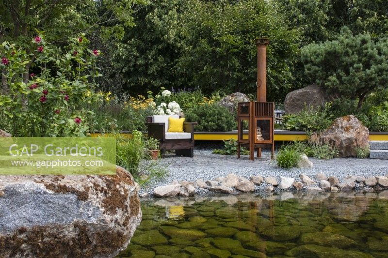 Une piscine avec bord de galets et gros rocher menant à un coin salon avec poêle à bois extérieur - Hurtigruten : The Relation-Ship Garden - designer Max Parker-Smith - RHS Hampton Court Flower Palace Garden Festival 2023. 