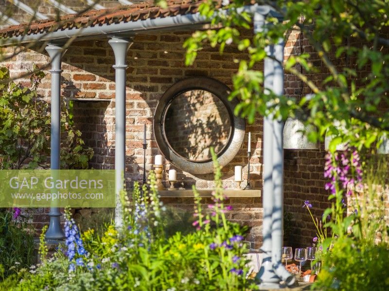Miroir circulaire sur mur de briques dans une salle à manger extérieure. The Savills Garden, Designer : Mark Gregory, RHS Chelsea Flower Show 2023, mai, printemps, été 
