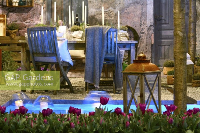 Contemporary Italian courtyard in front of the stone house with decorative table prepared for a spring evening party with candles, glasses and herbs in pot behind small pond with floating candles and row of purple tulips and lantern.