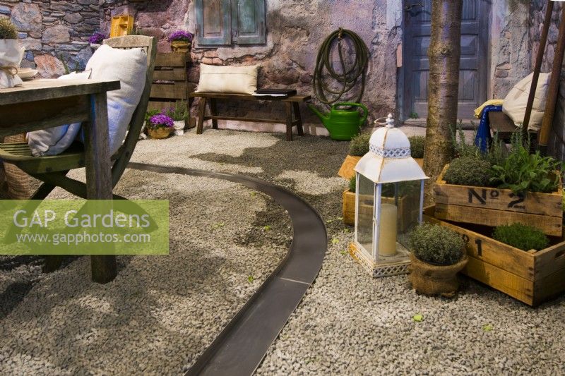Contemporary Italian courtyard in front of the stone house with courved water canal running on the gravel surface near herbs in pots and lantern.


