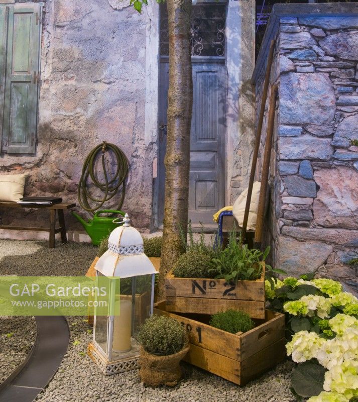 Exposition d'herbes et d'hortensias en pots et lanterne devant la maison en pierre de cour italienne contemporaine. Un canal d'eau qui coule sur la surface de gravier. 