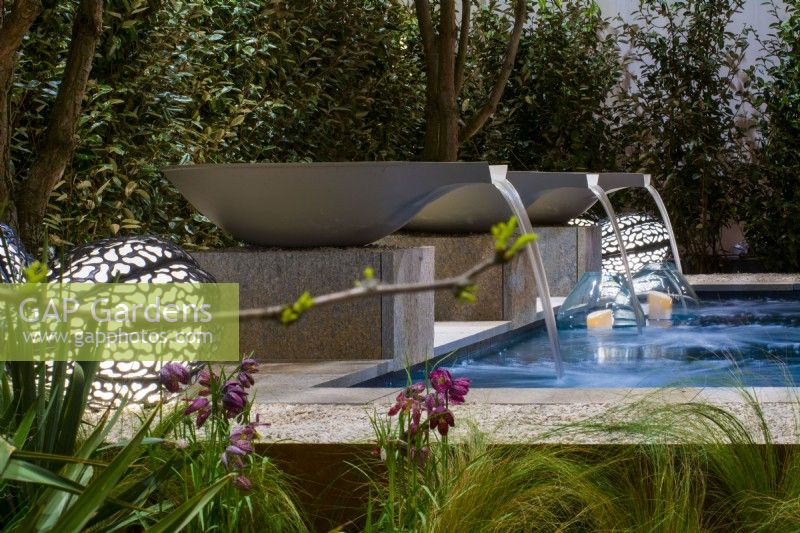 Water falls from three bowls into pool, nearby decorative floor lamps lit up the evening terrace. In foreground, Stipa and fritillaries. Designer: Vetschpartner, Berger Gartenbau and Livingdreams. Giardina-Zurich, Swiss.

