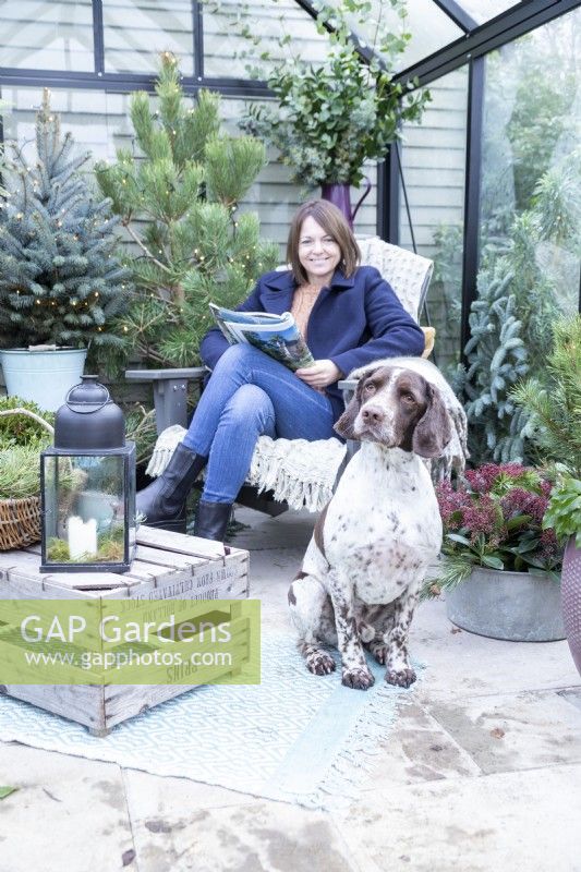 Dog sitting by woman on chair with magazine in inside decorated greenhouse