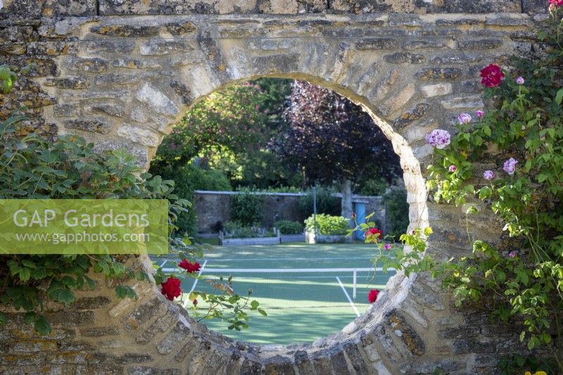 Une fenêtre lunaire dans un mur en pierre donnant sur un court de tennis, encadrée de Rosa 'Ferdinand Pichard' 
