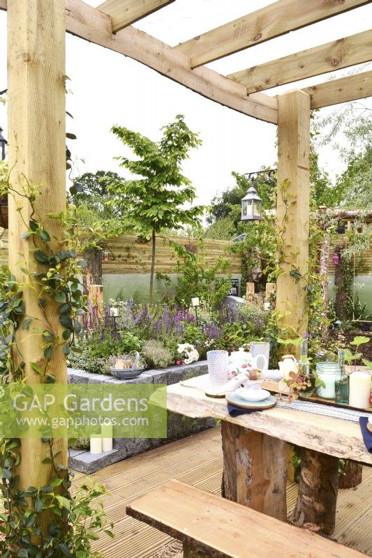 Pergola en bois avec un toit protecteur en plexiglas avec une table dressée dans un jardin d'inspiration boisée entouré d'une clôture en planches de bois. JuinConceptrice : Mary Anne Farenden. Bord Bia Bloom, Super Garden, Dublin, Irlande. 