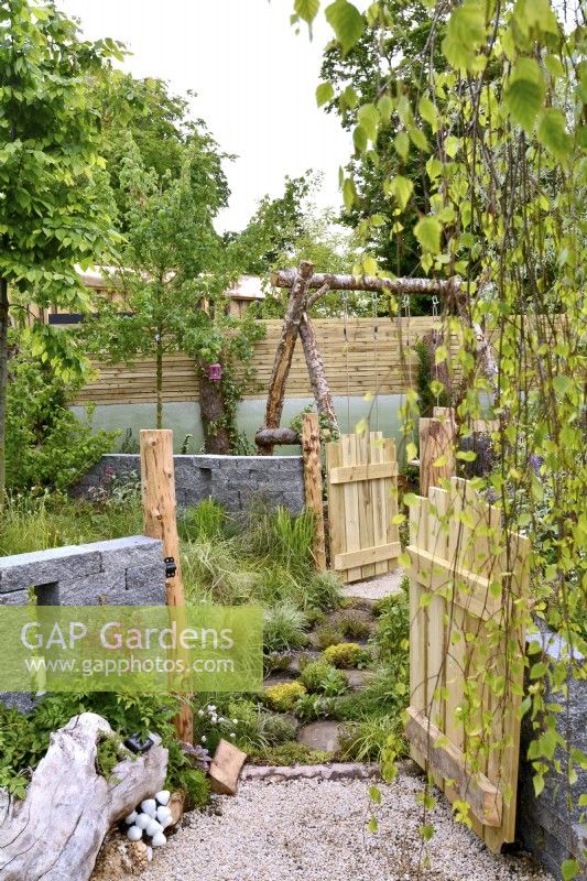 Opened wooden a gate leads into a grass area for dogs, alongside a large tree stump lying on a gravel surface used as a flowerbed and seating area next to a decorative Connemara wall system  in a woodland inspired garden. June
Designer: Mary Anne Farenden. Bord Bia Bloom, Super Garden, Dublin, Ireland.

