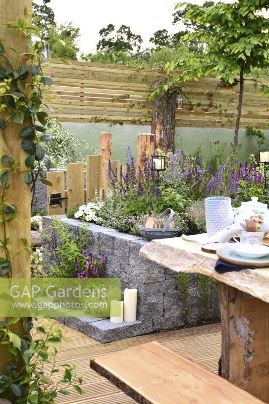 Rustic woodland inspired garden surrounded by a wooden planks fence with raised bed  made of Connemara decorative wall system. Planted with Thymus vulgaris, Leucanthemum superbum, Salvia Nemorosa, Sorbus aria. June
 


