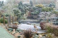 L'étang gelé et la terrasse en bois un matin d'hiver glacial. Pot de Prunus incisa 'Kojo-no-mai', ornements en bois de canard et poteaux en ardoise. Conifères sur rocaille au-delà.