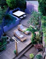 Vue sur jardin sur deux niveaux avec pergola avec vigne, chaises longues, terrasse en bois, cabanon, parterre de fleurs surélevé avec des herbes bleues, paillis de coquillages, Hosta et Acer en pots